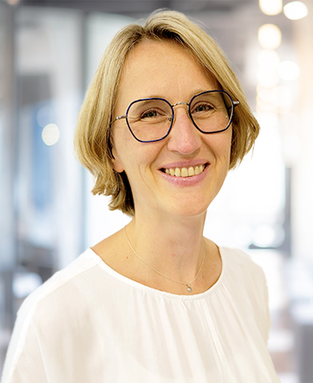 Eine lächelnde Frau mit Brille und blondem Haar trägt eine weiße Bluse.